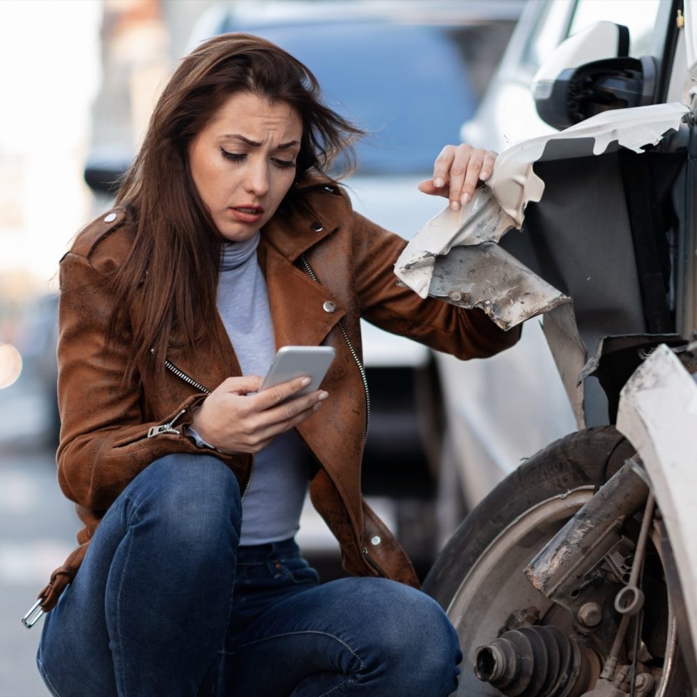 Young sad woman text messaging on smart for after a car crash on the road.